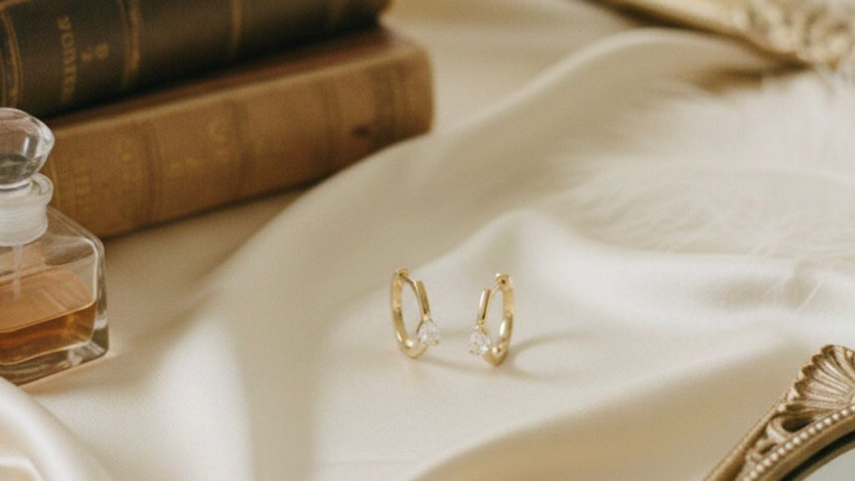 Gold hoop earrings on a white fabric surface with a small bottle of perfume and books in the background.
